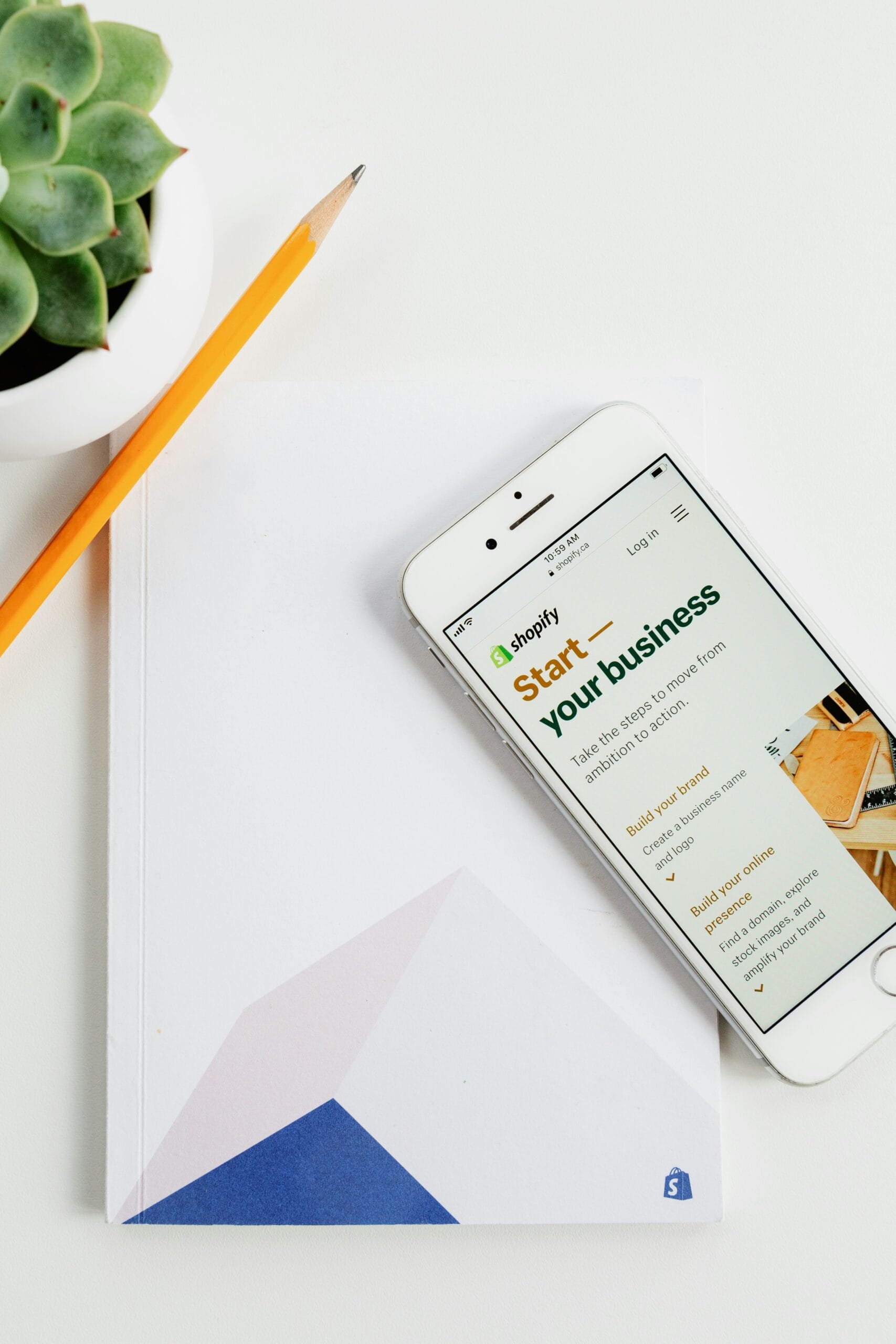 Un smartphone affichant la page d'accueil de Shopify, posé sur un carnet blanc avec un crayon et une plante en pot à côté, symbolisant le choix d'une plateforme e-commerce.