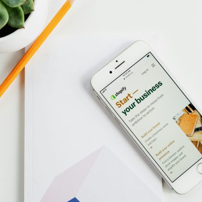 Un smartphone affichant la page d'accueil de Shopify, posé sur un carnet blanc avec un crayon et une plante en pot à côté, symbolisant le choix d'une plateforme e-commerce.